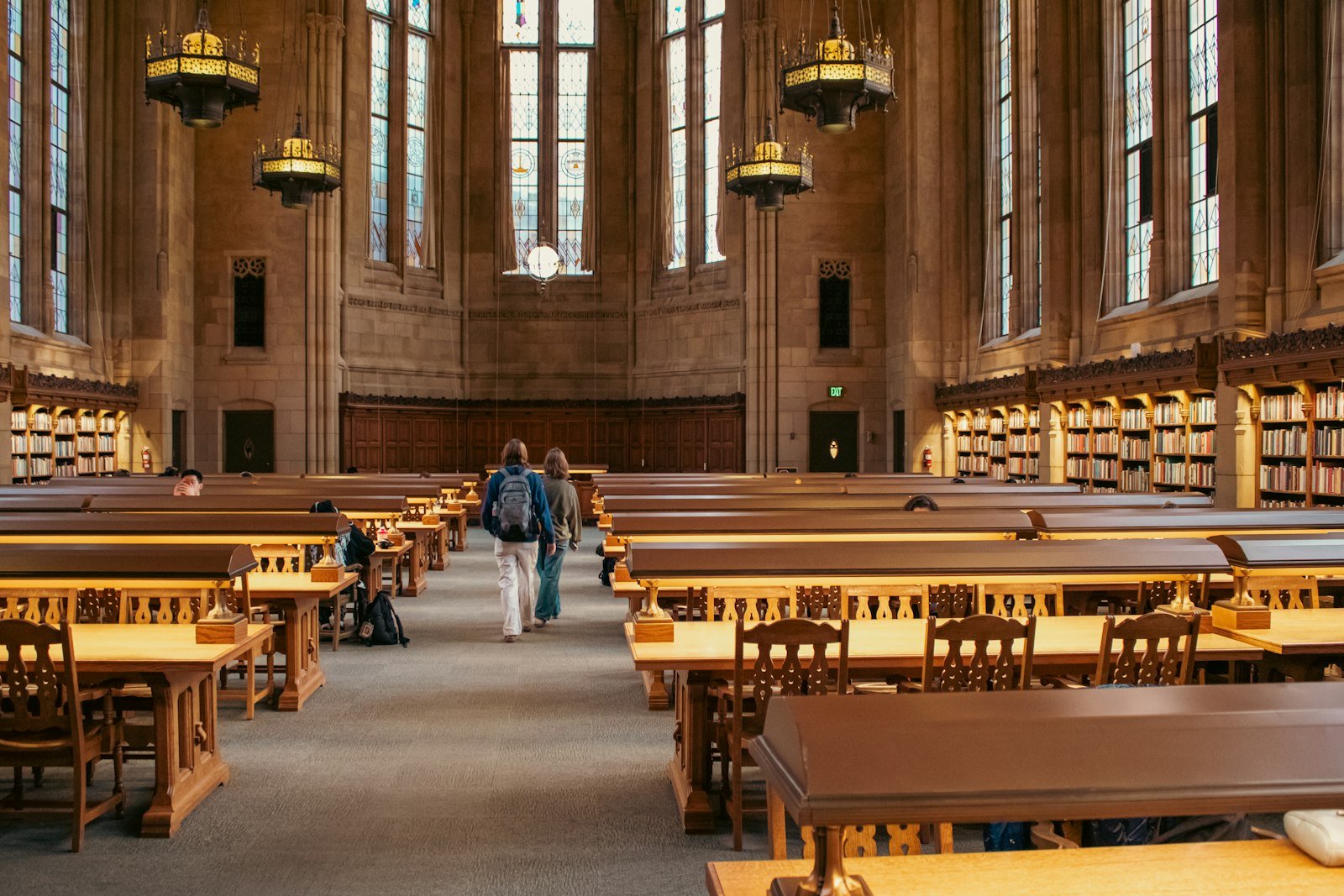 Students walking through a university library reading room lined with bookshelves